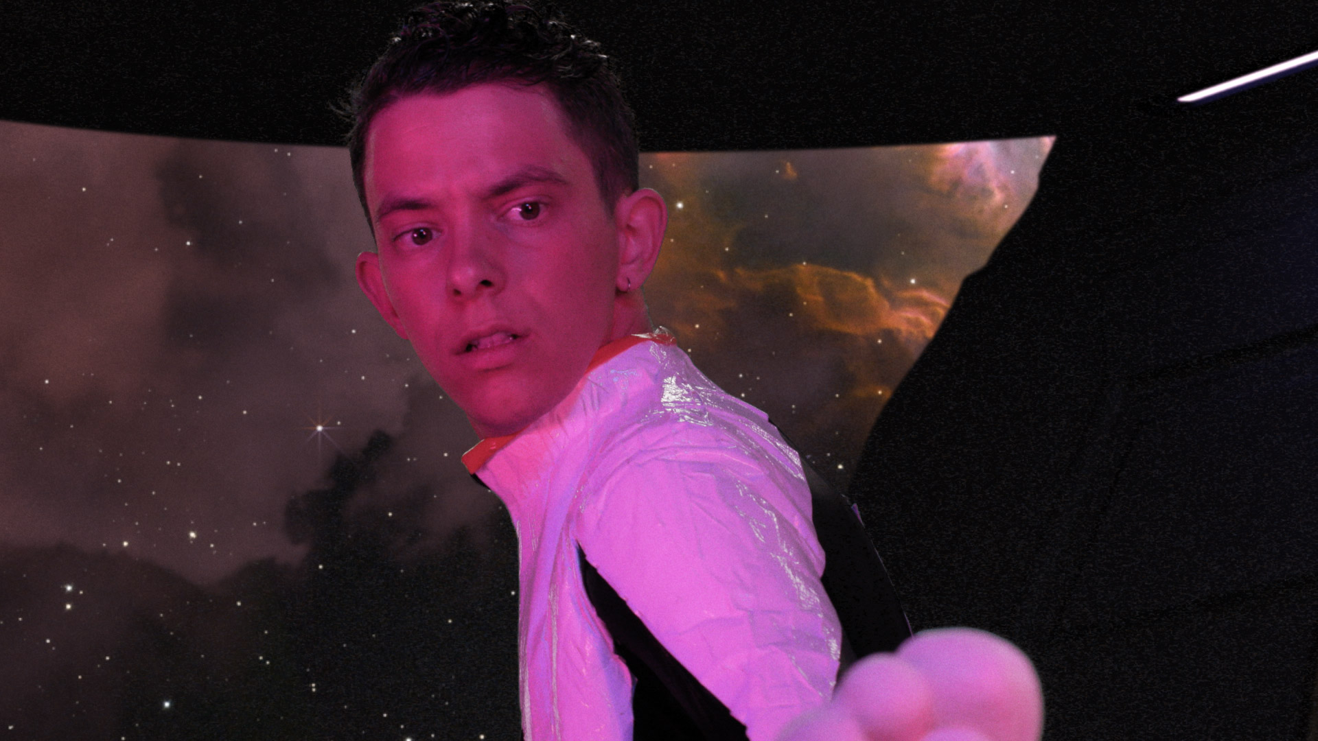 Film still of Mishaps in Spacetime: The steering joystick stands on the cuntpit’s counter. In front of it is the glass window, showing stars overlayed with a diffuse reflection of the joystick and the room, most noticeably monitors with bright colours.