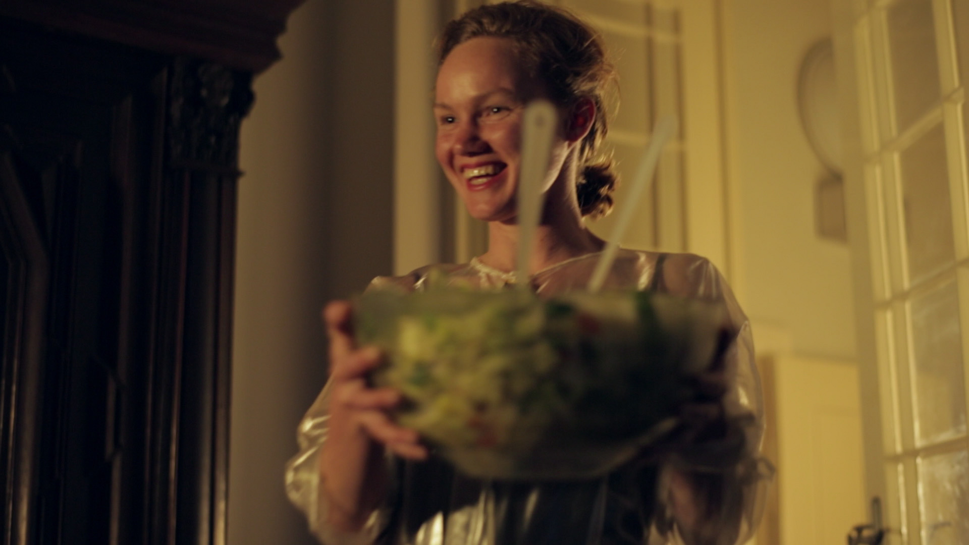 revolutionary road film still 2 - A medium shot from below of a housewife coming into a room, wearing a transparent suit and a wide grin, with a big bowl of salad in her hands.